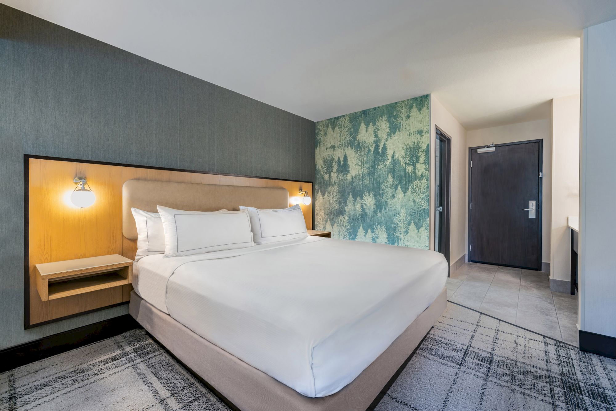 A modern hotel room features a large bed with white linens, bedside lamps, and forest-themed wallpaper near the entrance.