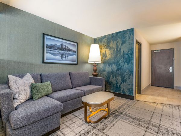 A modern living room with a gray sofa, decorative pillows, a framed landscape photo, a tall lamp, patterned wall, door, and coffee table.
