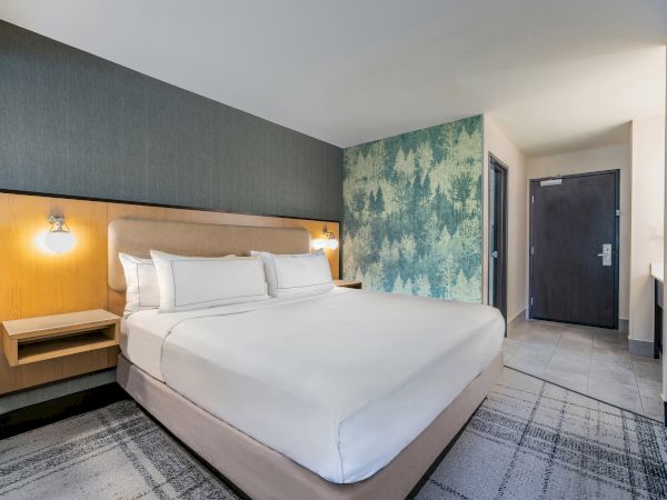 A modern hotel room with a large bed, stylish headboard, tree-patterned accent wall, and a door in the corner.