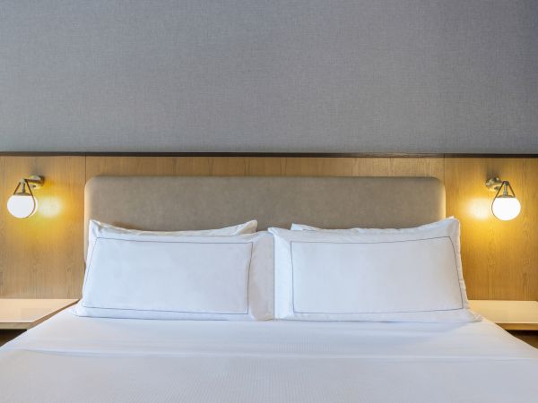A neatly made bed with white pillows and sheets, two bedside lamps are mounted on a wooden headboard.