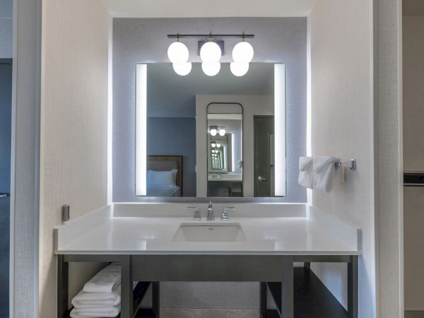 A modern bathroom vanity with a sink, illuminated mirror, and wall-mounted lights above, featuring neatly stacked towels underneath.