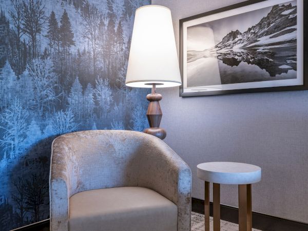 Room with a cozy chair, tall lamp, framed mountain photo, and blue forest-themed wallpaper.
