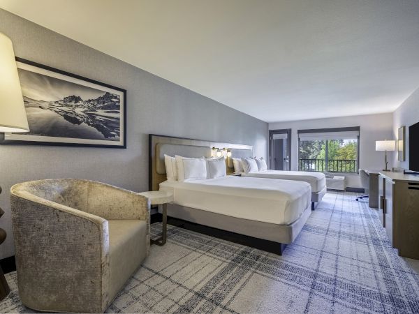 A modern hotel room with two beds, a sitting area, desk, TV, and large window overlooking greenery.