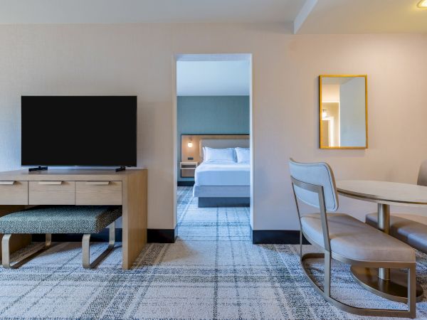 The image shows a hotel room with a TV, desk, chair, and a mirror, leading to a bedroom with a bed and lamp through an open doorway.