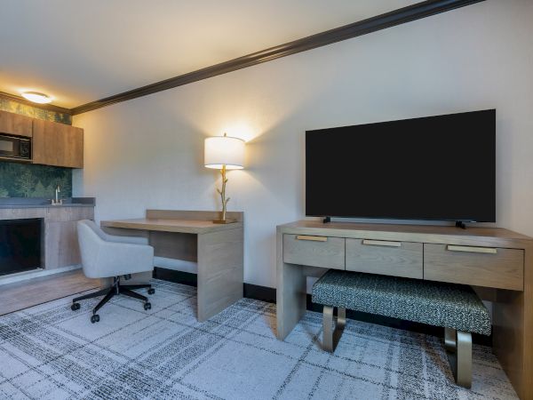The image shows a modern room with a desk, chair, lamp, microwave, mini fridge, and a flat-screen TV on a wooden cabinet.