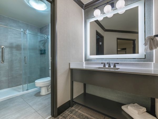 A modern bathroom with a glass shower, toilet, large mirror, a sink with lighting, and neatly folded towels on a shelf.
