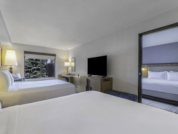 A modern hotel room with two beds, desk, TV, and window view, connected to another room with a bed through an open doorway.