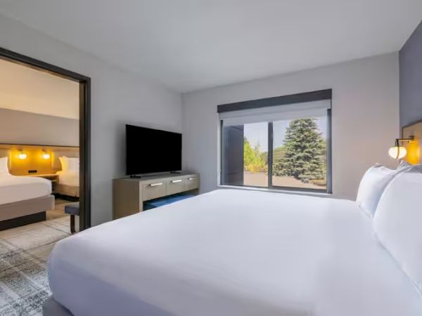 The image shows a hotel room with a bed, TV, window with a view, and an adjoining room visible through an open door.