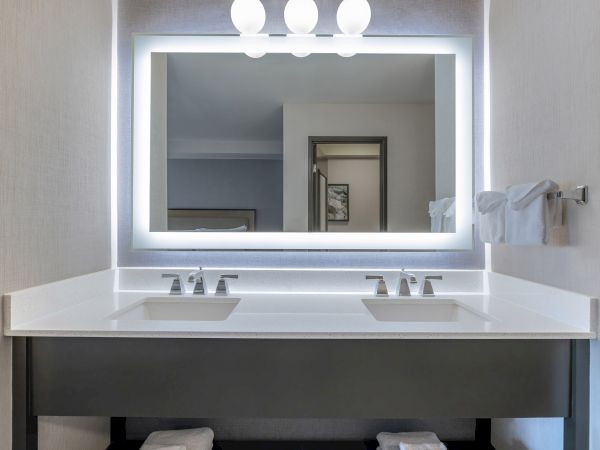 A modern bathroom with two sinks, a large illuminated mirror, and neatly folded towels on a towel rack in a hotel-themed setting.