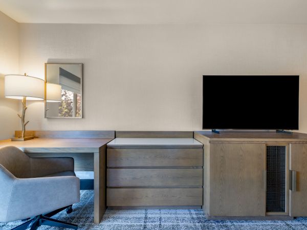 The image shows a modern office setup with a desk, chair, lamp, and a flat-screen TV on a wooden cabinet, in a bright room.